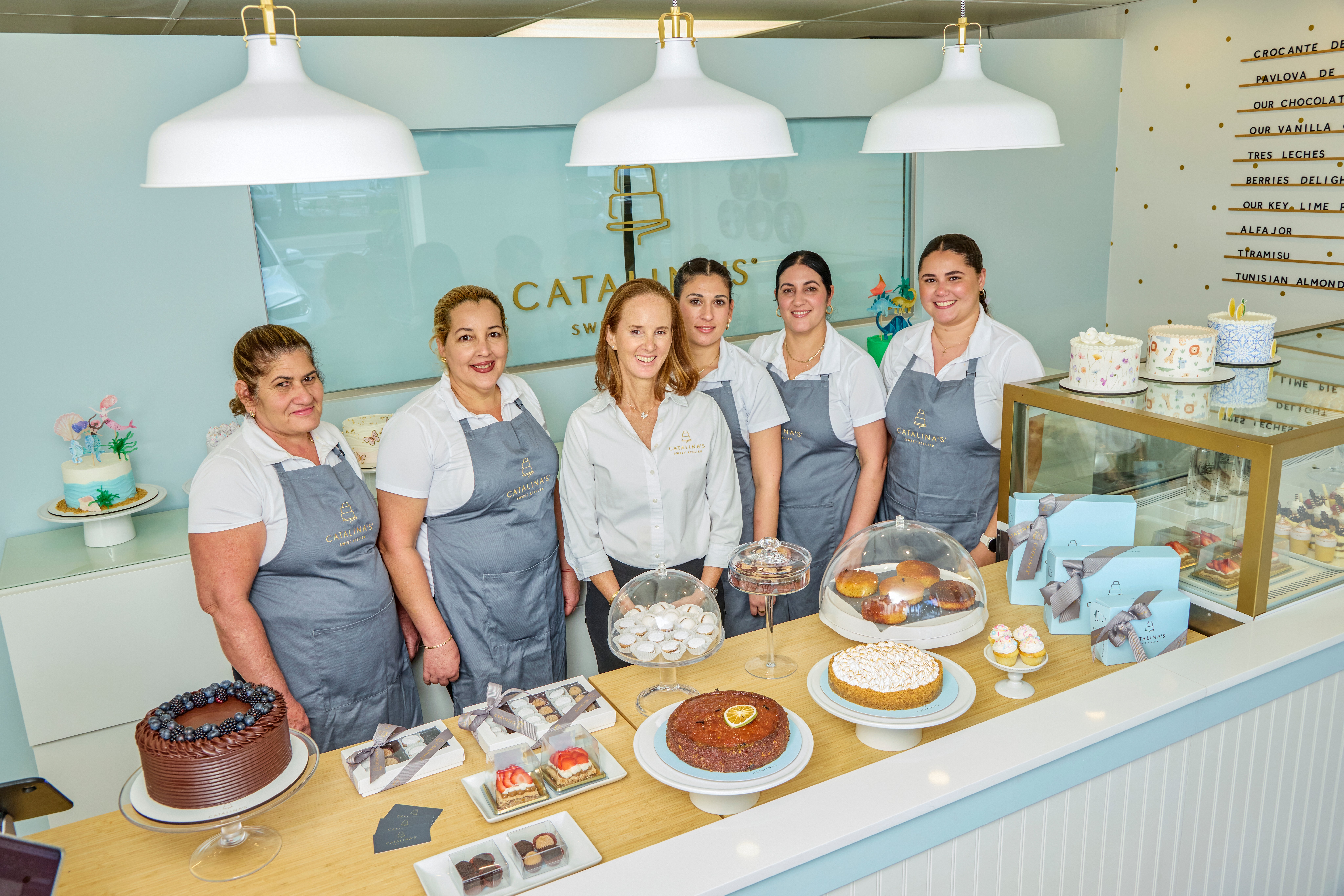 Six women in uniforms behind a bakery counter with cakes and baked goods, smiling at the camera.