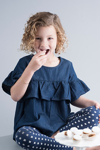 Young girl with curly hair sitting cross-legged, smiling, wearing a blue ruffled top and polka dot leggings, holding a cookie.