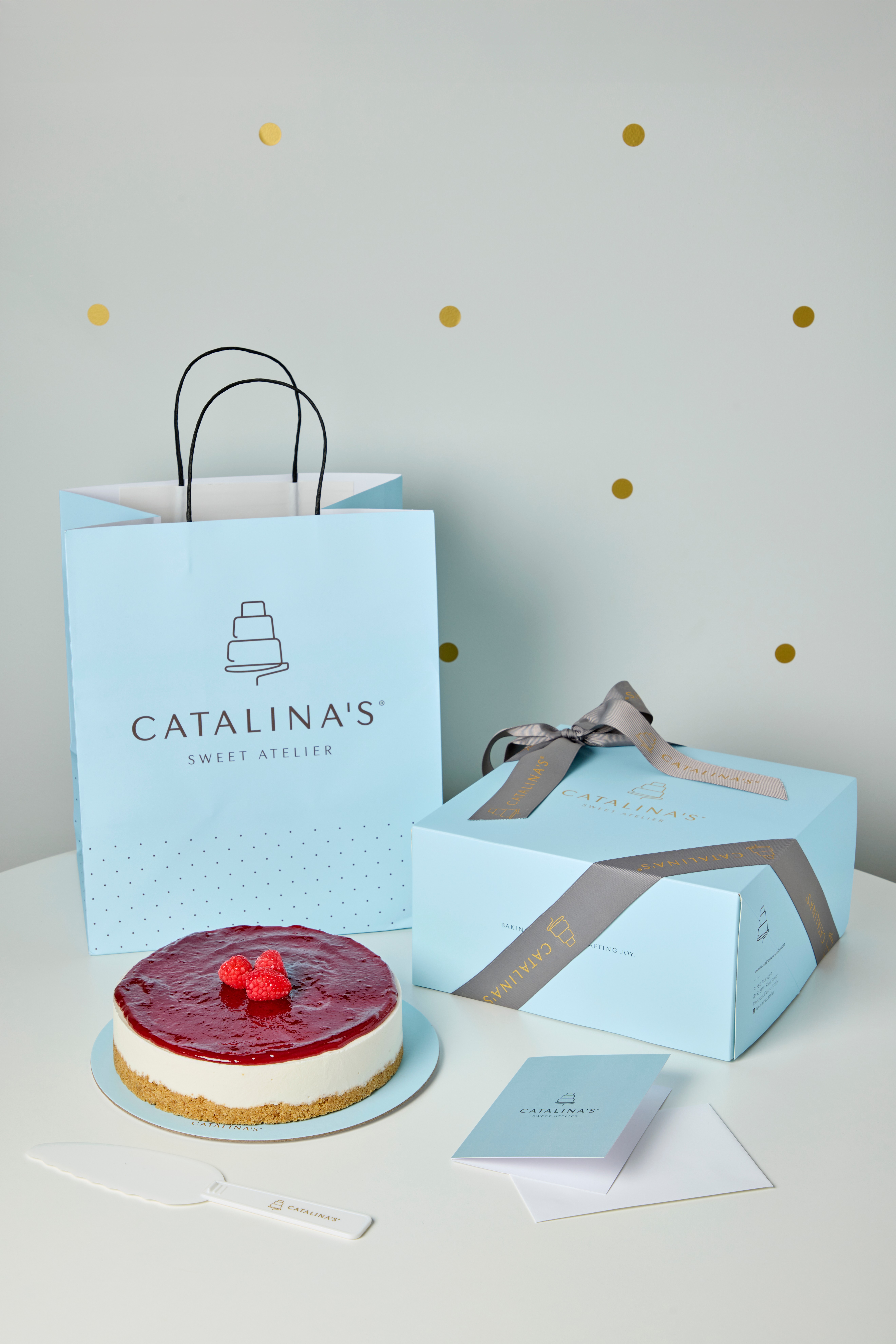 Cake with red fruit topping, gift box with ribbon, shopping bag, and cards on a white surface, decorated with gold dots.