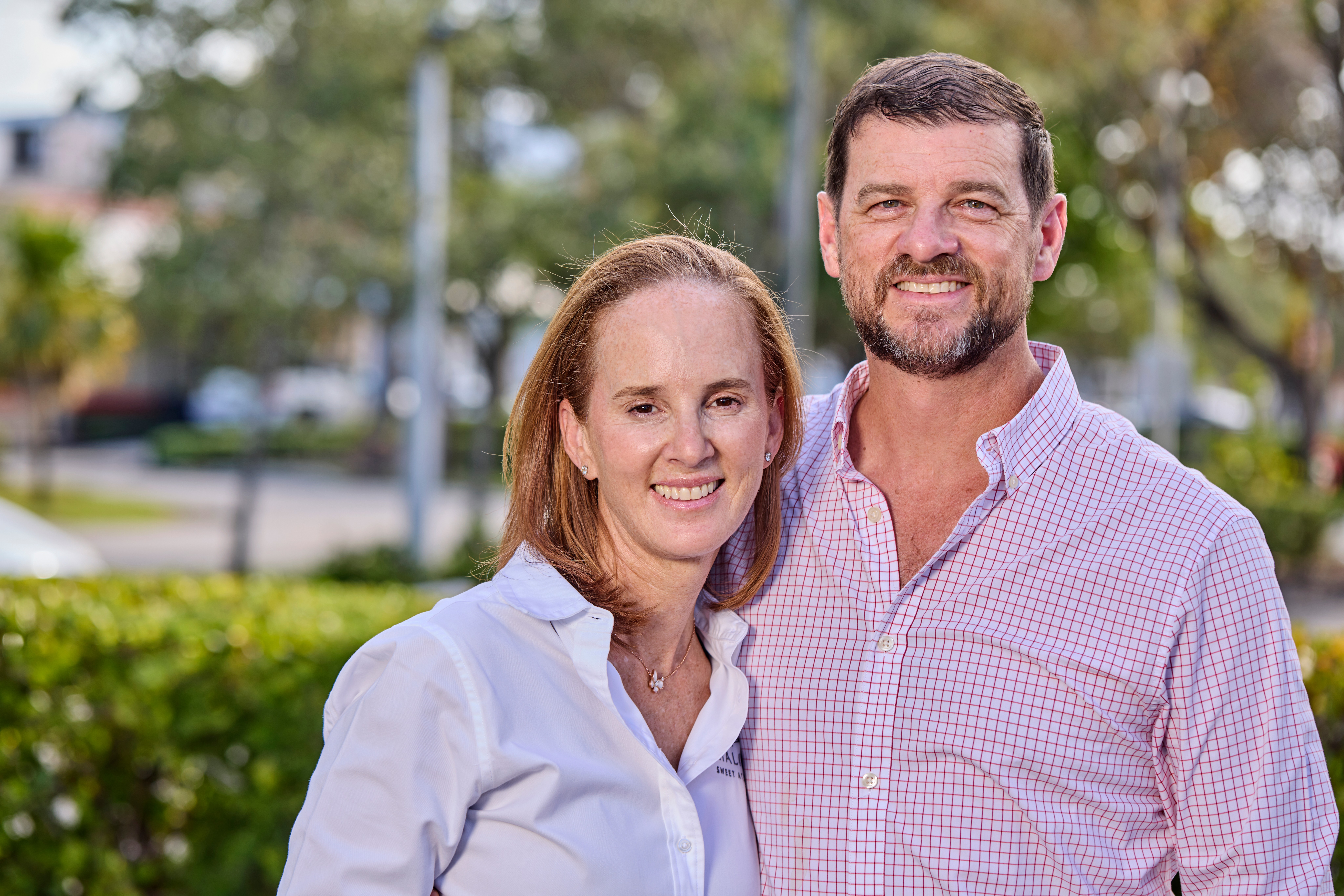 A smiling couple standing outdoors with trees and greenery in the background.