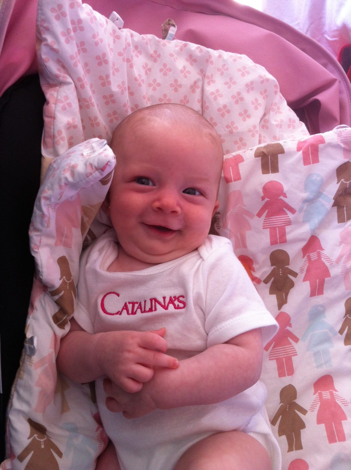 Baby lying on a patterned blanket, smiling, wearing a white shirt with red text, surrounded by pink and brown decorations.