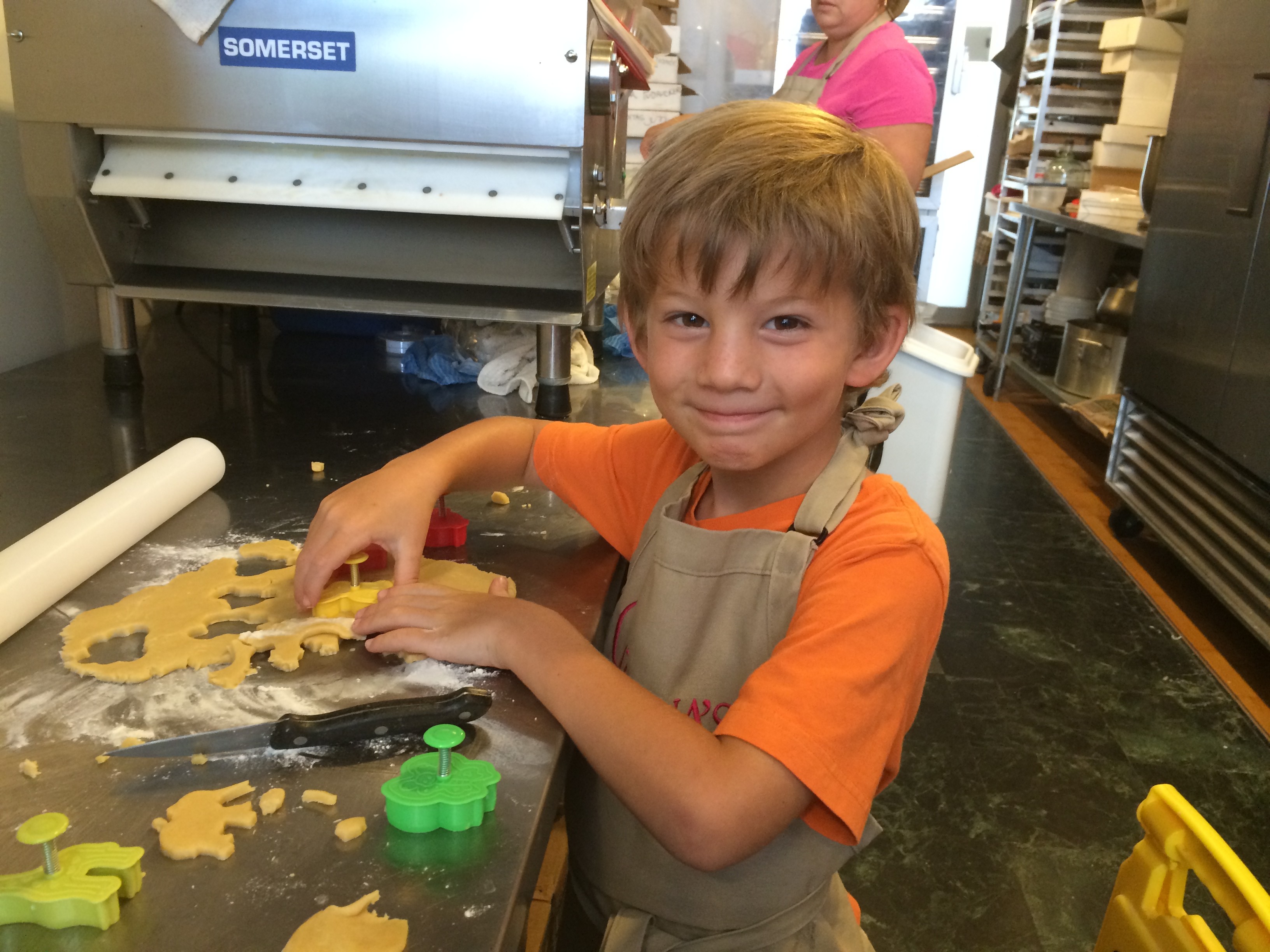 Young boy with a smile, wearing an orange shirt and apron, working with cookie dough in a bakery or kitchen.