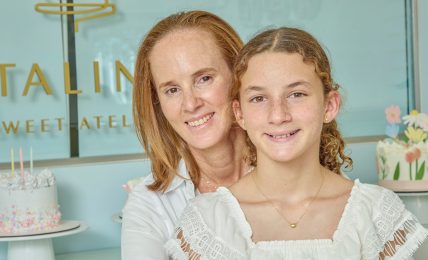 Two females smiling, one adult and one child, in front of a decorated cake and pastel background.