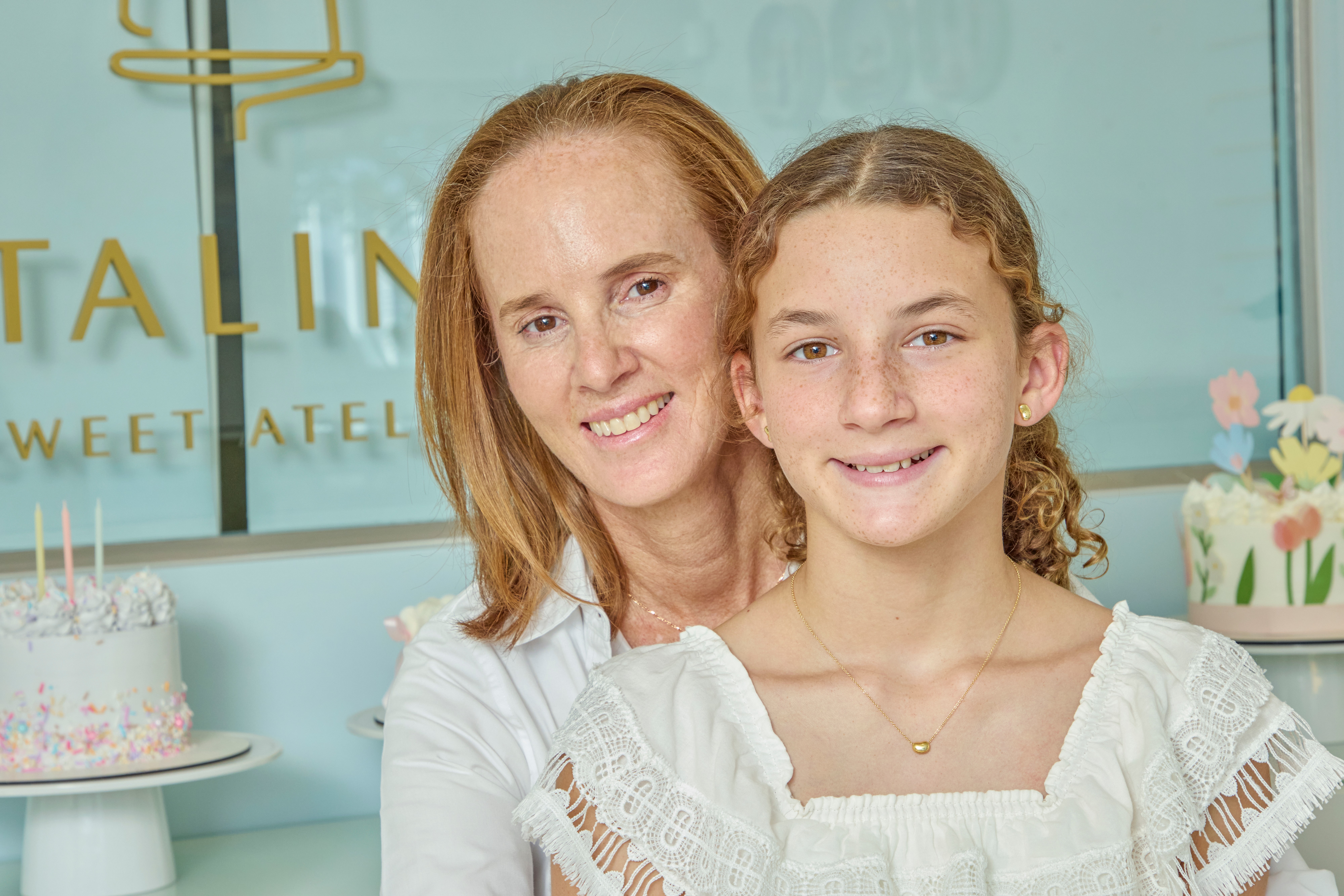 Two females smiling, one adult and one child, in front of a decorated cake and pastel background.